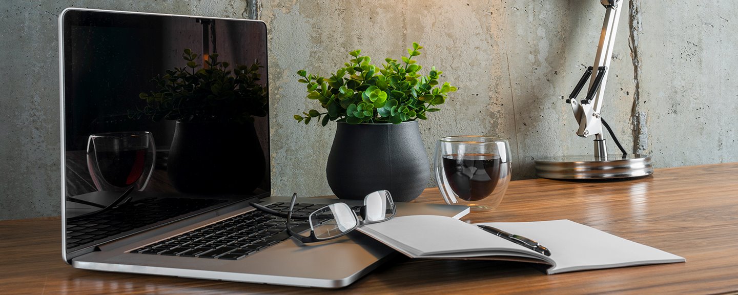 En laptop, ett par glasögon och ett öppet anteckningsblock på ett skrivbord i hemmamiljö