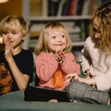 Glad flicka med Downs syndrom och storebror som klappar händerna, medan äldre syster visar något på surfplatta.