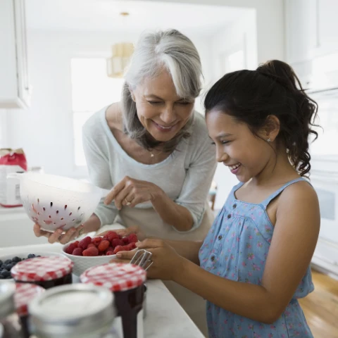 En äldre kvinna hjälper en ung flicka med brunt lockigt hår att koka jordgubbssylt